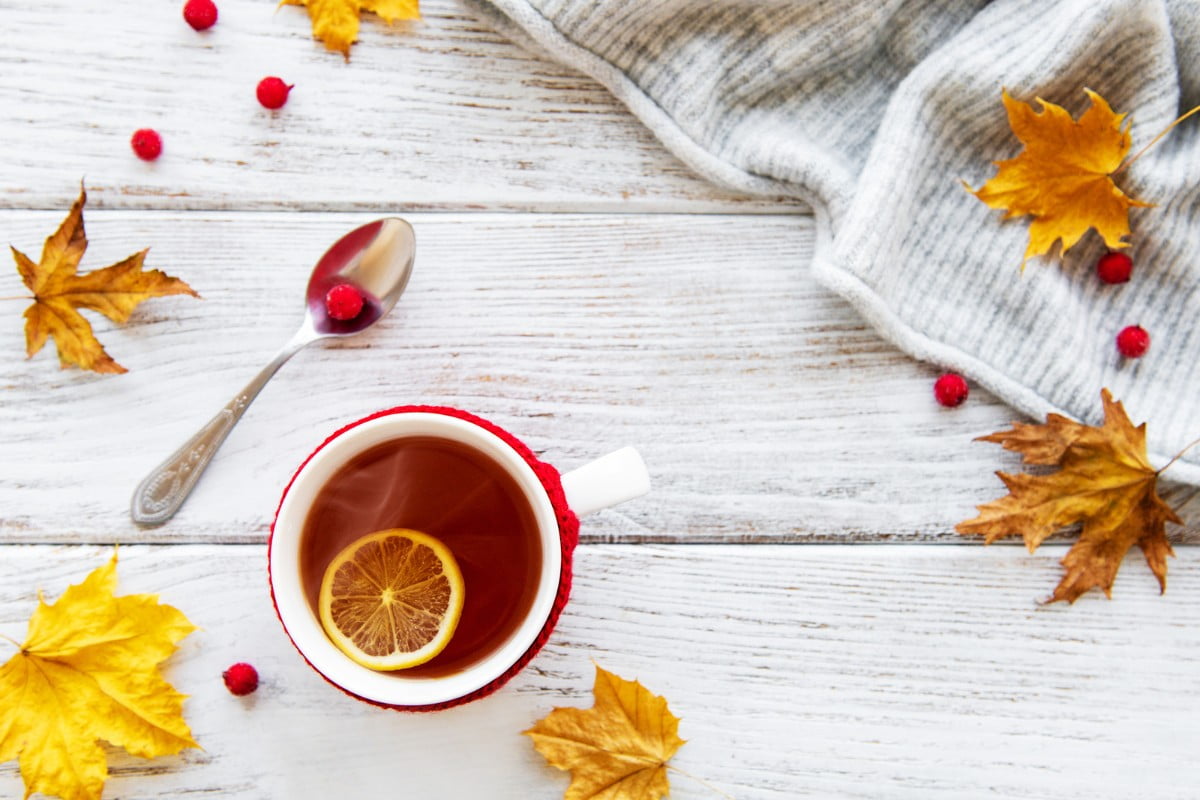 A mug of tea with sliced lemon on a white wooden surface surrounded by autumn leaves.